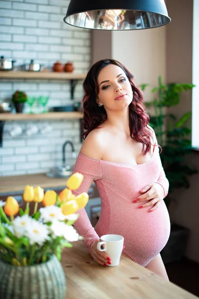 Young pregnant woman in pink bodysuit drinking water in the kitchen. How to get rid of morning sickness and toxemia. Drugs and vitamins for pregnancy — ストック写真