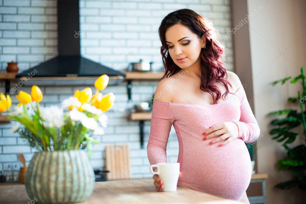 Young pregnant woman in pink bodysuit drinking water in the kitchen. How to get rid of morning sickness and toxemia. Drugs and vitamins for pregnancy