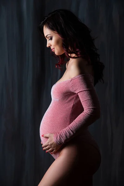 Vacker silhuett av en gravid kvinna på en mörk bakgrund. En brunett flicka i en rosa kropp passar varsamt omfamnar en gravid mage — Stockfoto