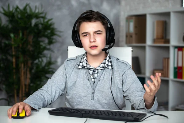 A teenager boy in headphones playing computer games at pc at home in the interior in front of a computer monitor