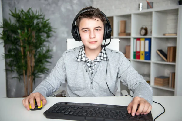 A teenager boy in headphones playing computer games at pc at home in the interior in front of a computer monitor — Stock Photo, Image