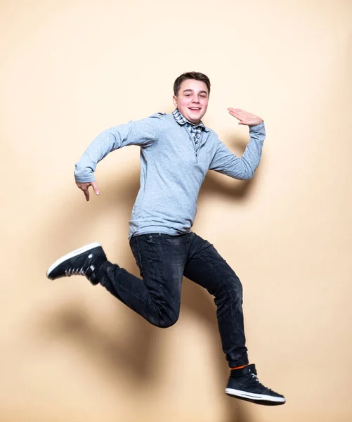 Mid-Air shot van vrolijke, succesvolle, gelukkige tiener kerel in Casual outfit springen met glimlach, handen omhoog, triomfantelijke, gebaren tegen beige achtergrond — Stockfoto