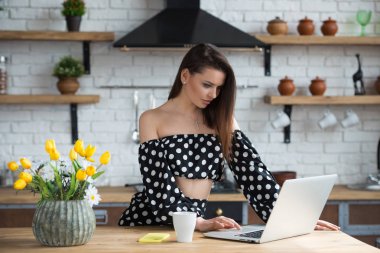 Ahşap masada dizüstü bilgisayar ile rahat mutfakta oturan bir polka nokta elbise çekici esmer kız blogger