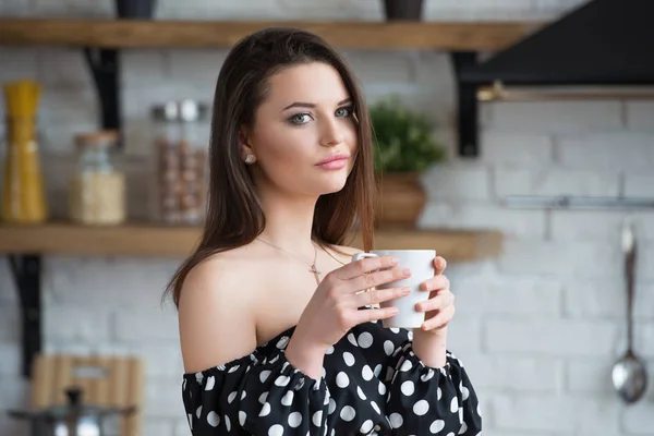 Atrakcyjna dziewczyna uśmiechnięta w polka dot sukienka pije kawę i siedzi na drewnianym stole w przytulnej kuchni rano. Przerwa na kawę — Zdjęcie stockowe