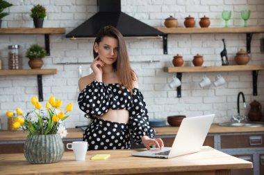 Çekici esmer kız blogger bir polka nokta elbise rahat mutfakta oturan ve ahşap masada dizüstü bilgisayarda haber okuma