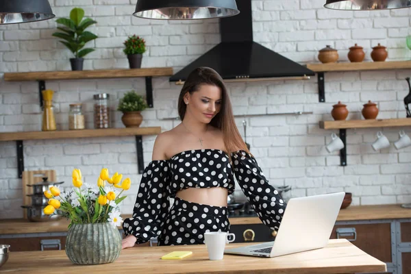 Attractive brunette girl blogger in a polka dot dress sitting in the cozy kitchen, drinking coffee tea and reading news on laptop at the wooden table — Stock Photo, Image