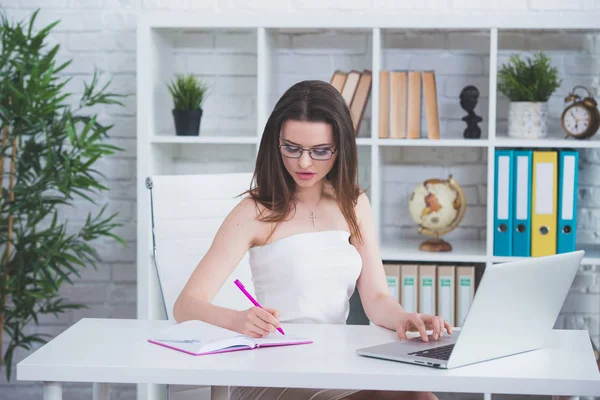 Młoda Dziewczyna Brunetka w białej sukni siedzi w biurze przy stole i pisze ważne notatki w swoim notebooku. Kobieta pracuje na laptopie — Zdjęcie stockowe