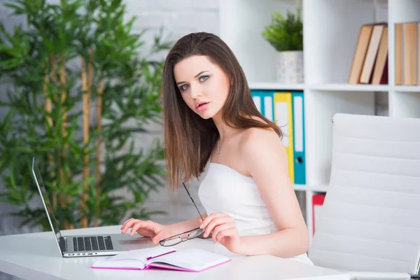 En ung brunett flicka i en vit klänning sitter på kontoret vid bordet. Kvinna arbetar på den bärbara datorn — Stockfoto