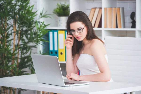 En ung brunett flicka med glasögon i en vit klänning sitter på kontoret vid bordet. Kvinnan jobbar på laptopen. Låg syn eller mode tillbehör — Stockfoto