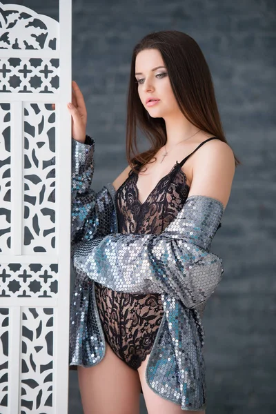 Beautiful sexy brunette girl in a black lace jumpsuit and silver jacket stands near the white openwork wooden screen — ストック写真