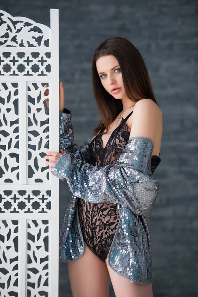 Beautiful sexy brunette girl in a black lace jumpsuit and silver jacket stands near the white openwork wooden screen — ストック写真