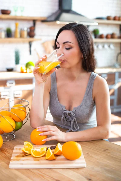 Lächelnde junge Frau im grauen T-Shirt trinkt frischen Orangensaft in der Küche — Stockfoto