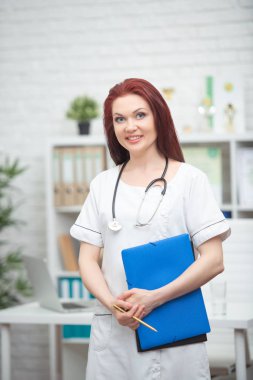 Bir stetoskop ile üniformalı kadın doktor gülümseyen ve elinde mavi bir klasör onun tıbbi ofiste ayakta ve hastalar karşılar