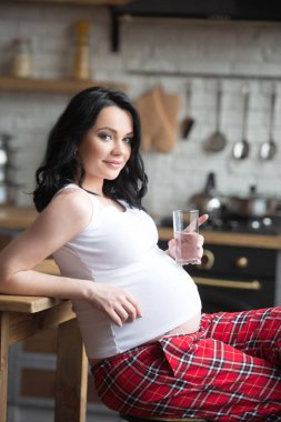 Damalı pijamalarıyla genç ve güzel bir kadın mutfakta bir bardak saf su içiyor. Güzel sağlıklı gebelik ve taze cilt