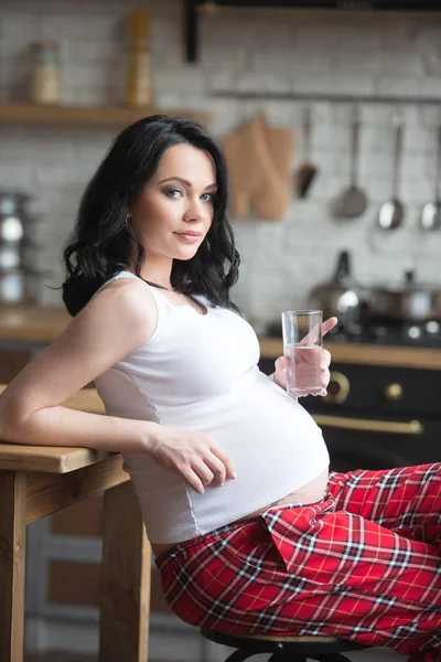 チェッカー付きパジャマの若い美しい女性が台所で純水を飲んでいます。美しい健康な妊娠と新鮮な肌 — ストック写真