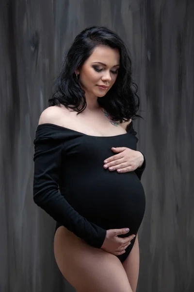 Una hermosa joven morena embarazada con un mono negro está abrazando su barriga. Embarazo con estilo, salud de la mujer, maternidad feliz — Foto de Stock