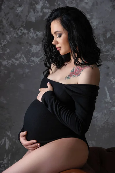Uma bela jovem mulher morena grávida em um macacão preto está abraçando sua barriga. Gravidez elegante, saúde da mulher, maternidade feliz — Fotografia de Stock