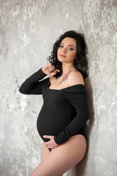 Uma bela jovem mulher morena grávida em um macacão preto está abraçando sua barriga. Gravidez elegante, saúde da mulher, maternidade feliz — Fotografia de Stock