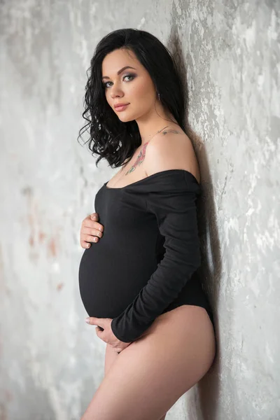 Una hermosa joven morena embarazada con un mono negro está abrazando su barriga. Embarazo con estilo, salud de la mujer, maternidad feliz — Foto de Stock