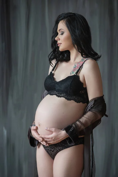 O conceito de gravidez. Mulher grávida bonita em roupa interior preta abraçando sua barriga contra um fundo escuro — Fotografia de Stock