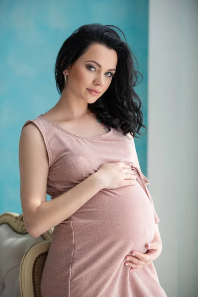 Salud de la mujer y embarazo. Hermosa mujer embarazada en un vestido rosa suave está de pie junto a la ventana y abrazando su barriga — Foto de Stock