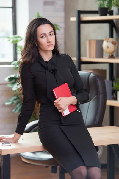 Beautiful stylish business woman with a red notebook in hand is standing in the office. Women's business and career — Stock Photo, Image
