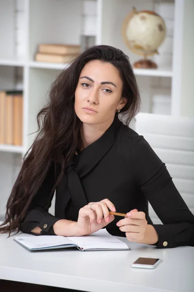 Brunetka dziewczyna w biznesie czarna sukienka siedzi przy stole w biurze i ciężko pracuje. Student pisze notatki, bierze notatki w Notatniku — Zdjęcie stockowe