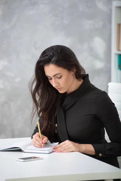 A brunette girl in a business black dress is sitting at a table in the office and is working hard. Student writes notes, takes notes in a notebook — Stock Photo, Image