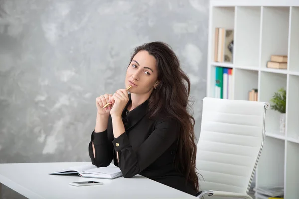 A brunette girl in a business black dress is sitting at a table in the office and is working hard. Student writes notes, takes notes in a notebook — Stock Photo, Image