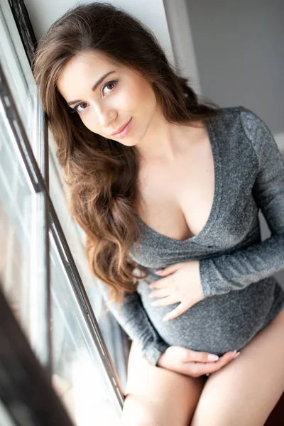 Красивая задумчивая беременная женщина с длинными вьющимися волосами сидит у окна в интерьере комнаты — стоковое фото