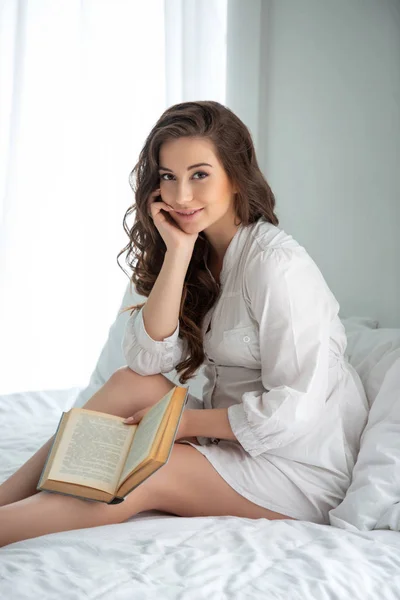 Mujer embarazada hermosa con el pelo ondulado largo leyendo un libro de la revista sentado en una cama en una habitación blanca soleada brillante —  Fotos de Stock