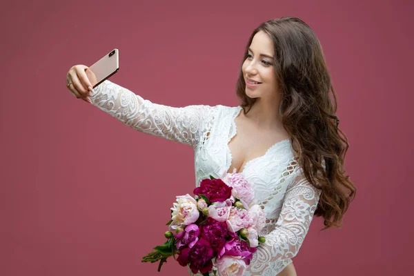Beautiful pregnant woman in lace white lingerie body suit makes a selfie with a bouquet of flowers peony — Stock Photo, Image