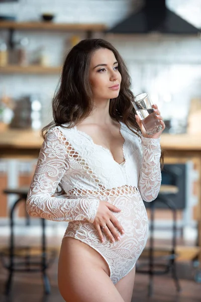 Vacker gravid kvinna med långt hår i en spets tröja dricka ett glas vatten i köket — Stockfoto