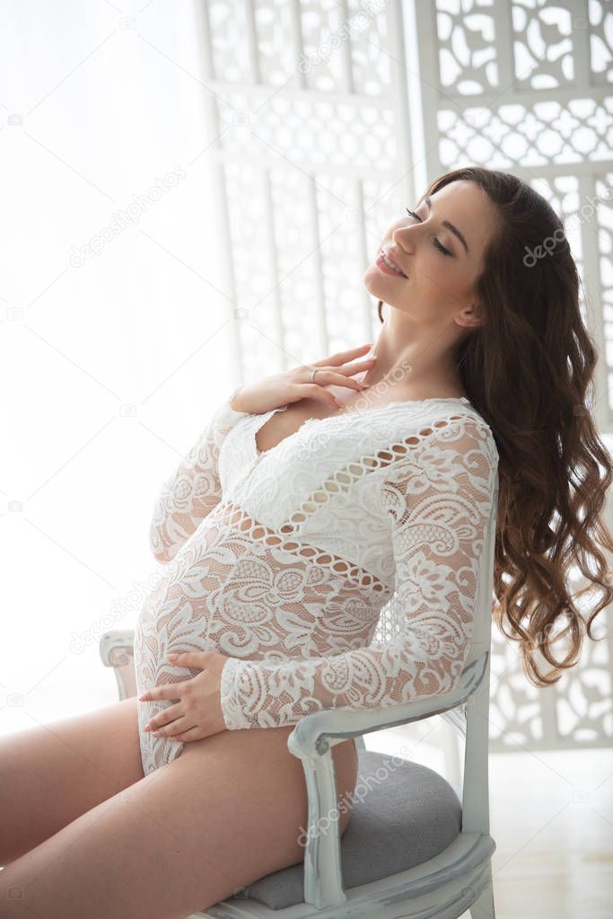 Beautiful pregnant woman with long wavy hair sitting on a chair, dressed in openwork lace white lingerie body suit