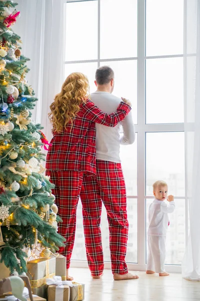 Szczęśliwa rodzina w piżamie Plaid w pobliżu dużego okna w salonie z choinki. Matka, ojciec i mały syn czekając na Świętego Mikołaja — Zdjęcie stockowe