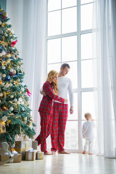 Szczęśliwa rodzina w piżamie Plaid w pobliżu dużego okna w salonie z choinki. Matka, ojciec i mały syn czekając na Świętego Mikołaja — Zdjęcie stockowe