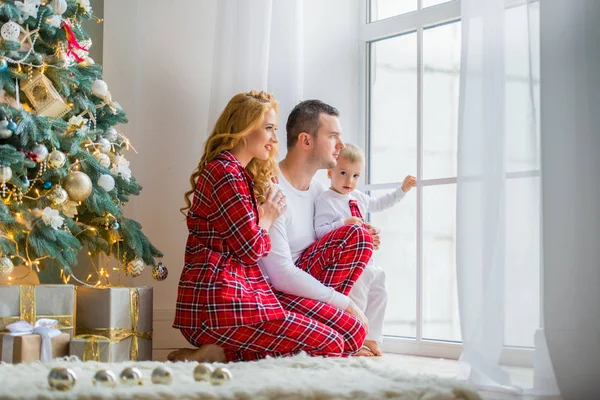 Szczęśliwa rodzina w piżamie Plaid w pobliżu okna w salonie z choinki. Matka, ojciec i mały syn czekając na Świętego Mikołaja — Zdjęcie stockowe