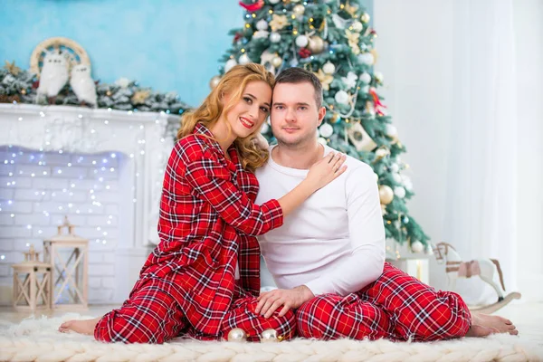 Любящие супруги в клетчатой пижаме сидят возле новогодней елки у камина и обнимаются. Любовь и счастливая семья — стоковое фото
