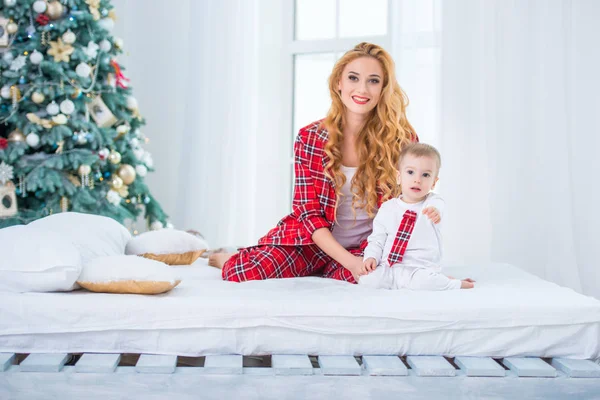Mom and son in checkered pajamas are sitting on the bed near the New Year tree and smiling