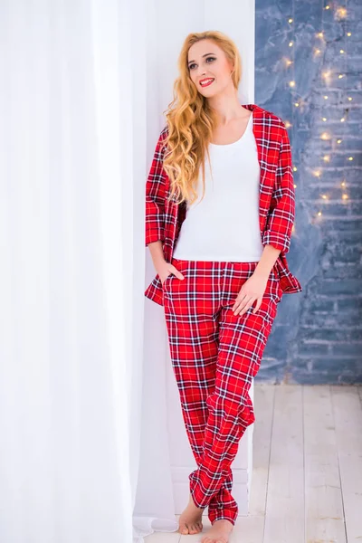 Vacker blond kvinna i rött rutig pyjamas står nära fönstret och visar bekväma hem kläder — Stockfoto