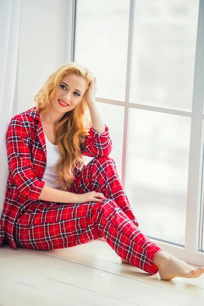 Piękna Blondynka w czerwonym piżamie w kratkę siedzi w pobliżu okna i pokazuje wygodne ubrania do domu — Zdjęcie stockowe