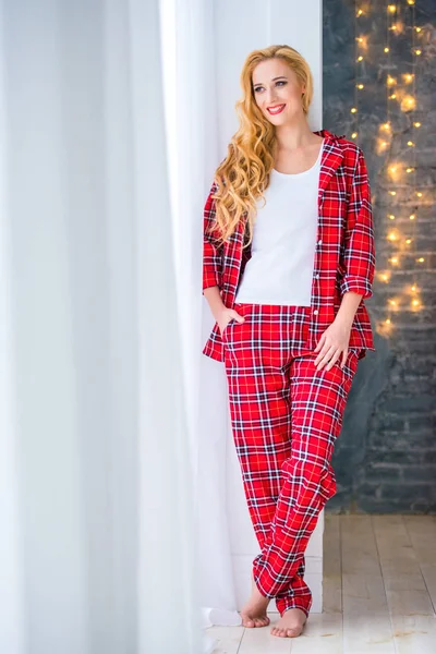 Gyönyörű szőke nő, piros kockás pizsama közelében áll az ablakon, és bizonyítja, kényelmes ruhákat otthon — Stock Fotó