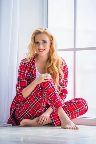 Gyönyörű szőke nő, piros kockás pizsama ül az ablak közelében, és bizonyítja, kényelmes ruhákat otthon — Stock Fotó