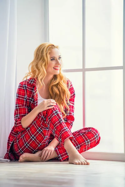 Schöne blonde Frau im rot karierten Pyjama sitzt am Fenster und demonstriert bequeme Heimkleidung — Stockfoto