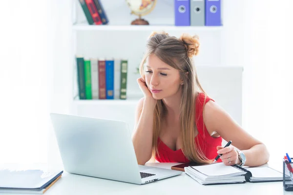 En ung kvinna i röd topp sitter på ett kontor vid ett datorbord. Kvinnliga företag — Stockfoto