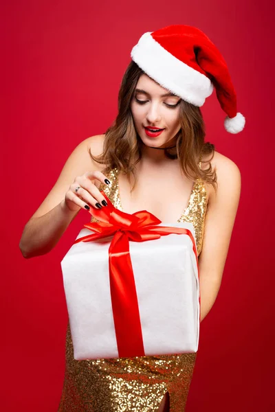 漂亮性感的圣诞老人，身穿金色优雅的晚礼服，头戴圣诞礼帽，手里拿着一份新年礼物。猜猜盒子里是什么？ — 图库照片