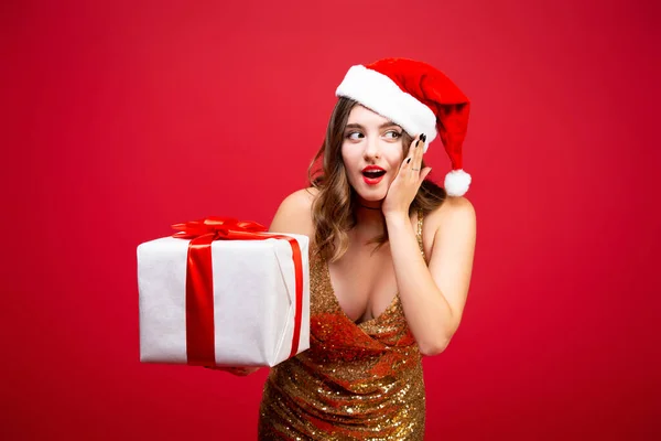 Piękna dziewczyna sexy Santa w złotej eleganckiej sukni wieczorowej i Boże Narodzenie kapelusz z prezentem na Nowy Rok w rękach. Emocje zaskoczenia na twarzy. Spróbuj zgadnąć. — Zdjęcie stockowe