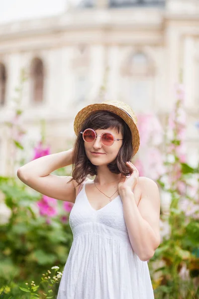 Portrait of a beautiful brunette in a white sundress, pink round glasses and a straw hat standing in a flower garden