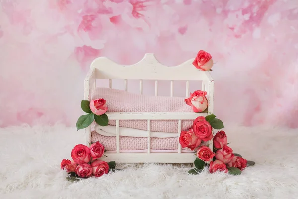 A white crib for a newborn baby adorned with pink rosebuds stands on a floral pink background and a white carpet