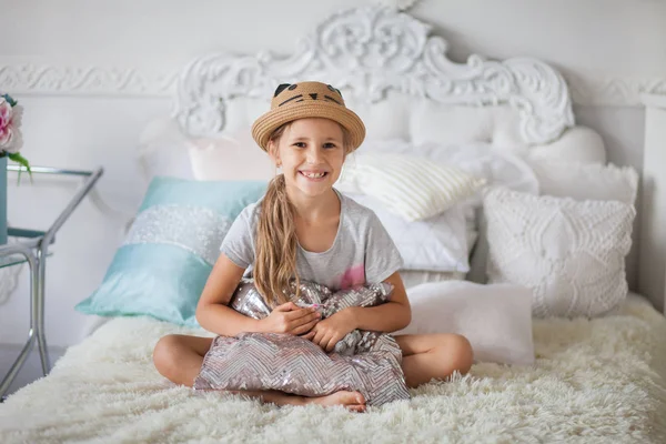 A girl in a hat sits on a bed in a white room, hugs a pillow, rest on the bed, furniture and children